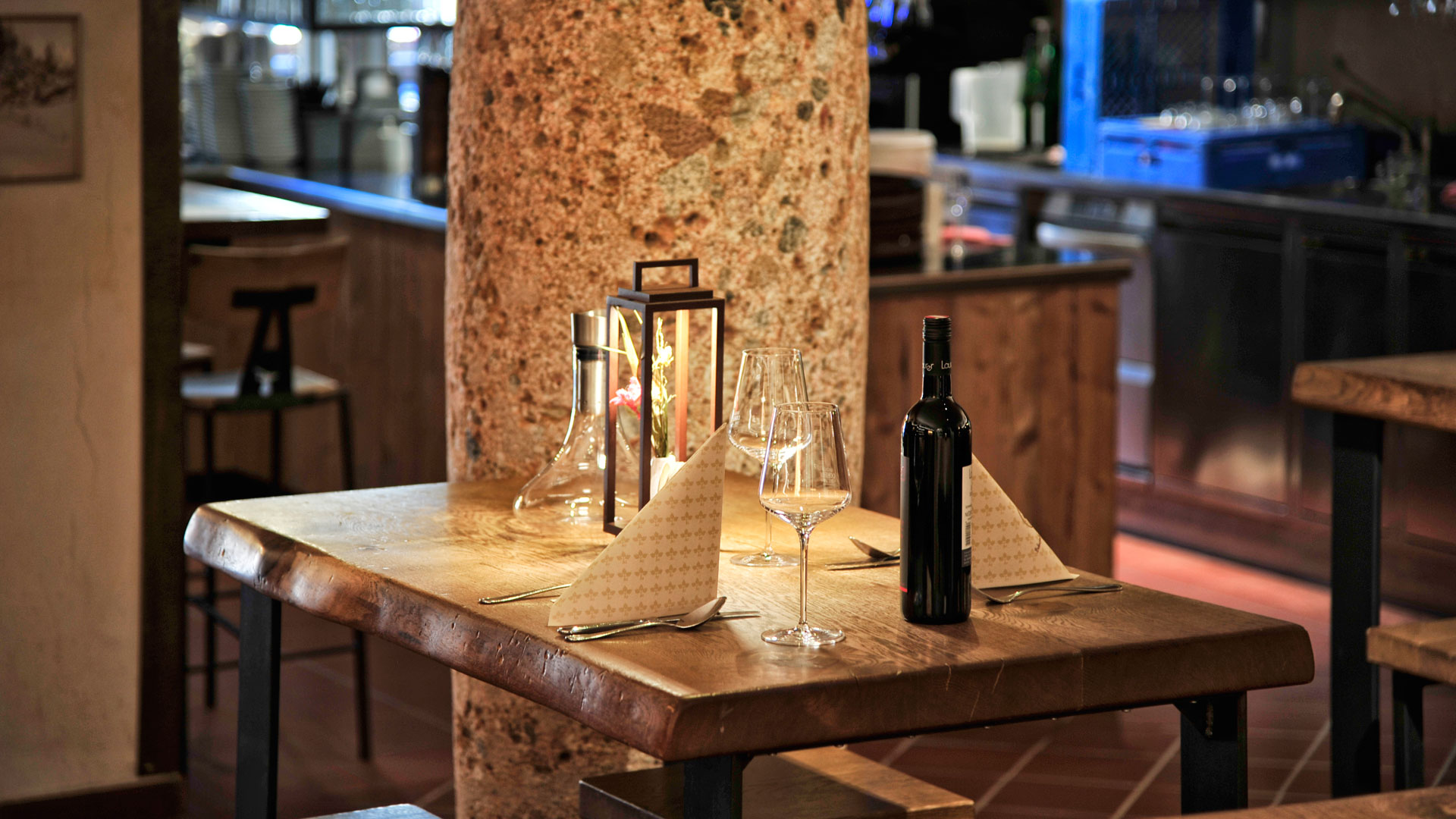 Stilvoll gedeckter Holztisch mit Wein, Gläsern und Laterne vor rustikalem Hintergrund mit Steinsäule und Bartheke.
