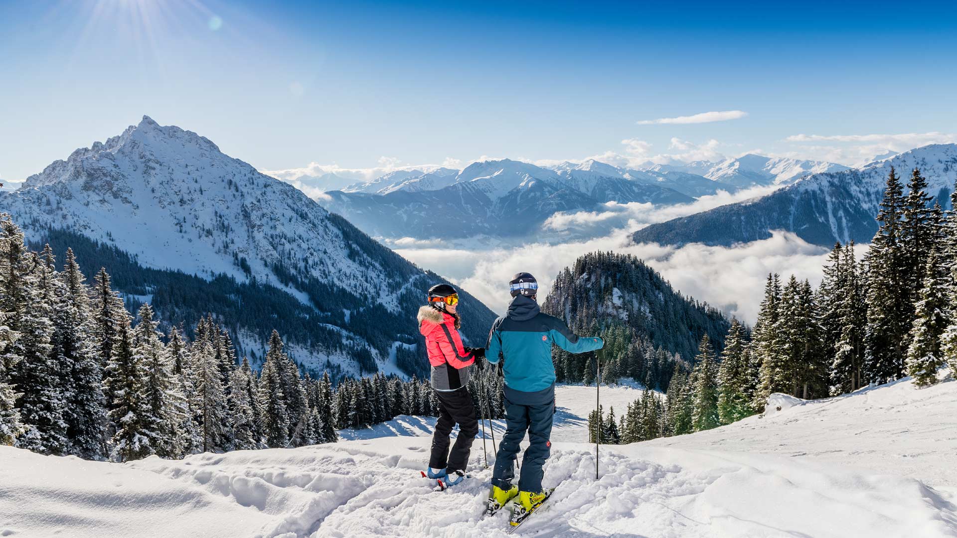 Zwei Skifahrer in Winterkleidung stehen am Pistenrand, blicken ins sonnige Bergtal mit schneebedecktem Wald und Gipfeln.